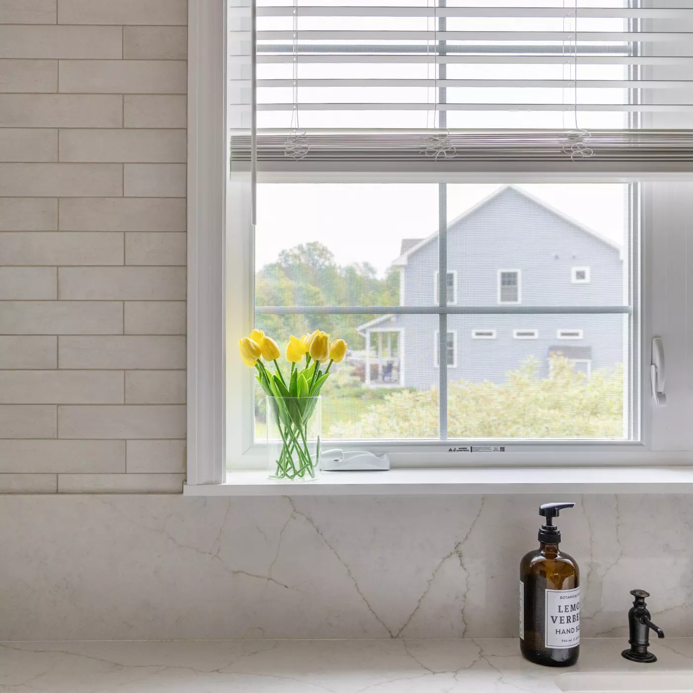 A kitchen window with yellow tulips and a view of a blue house outside.