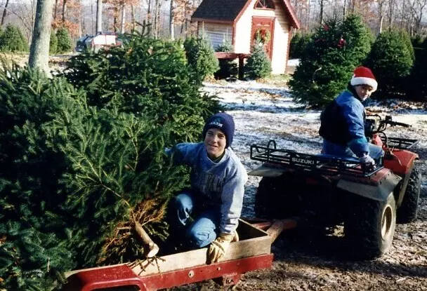 Two people at a Christmas tree farm &mdash; one kneels by a cut tree on a trailer, the other rides a red ATV in a Santa hat.