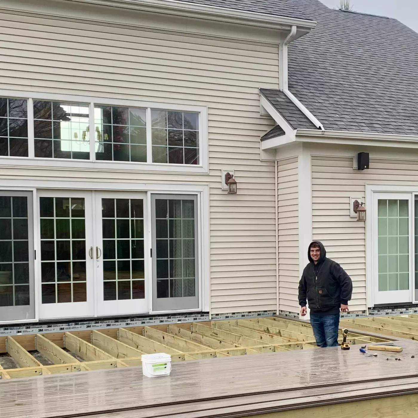 Person standing on a wooden deck frame in front of a house with large windows.