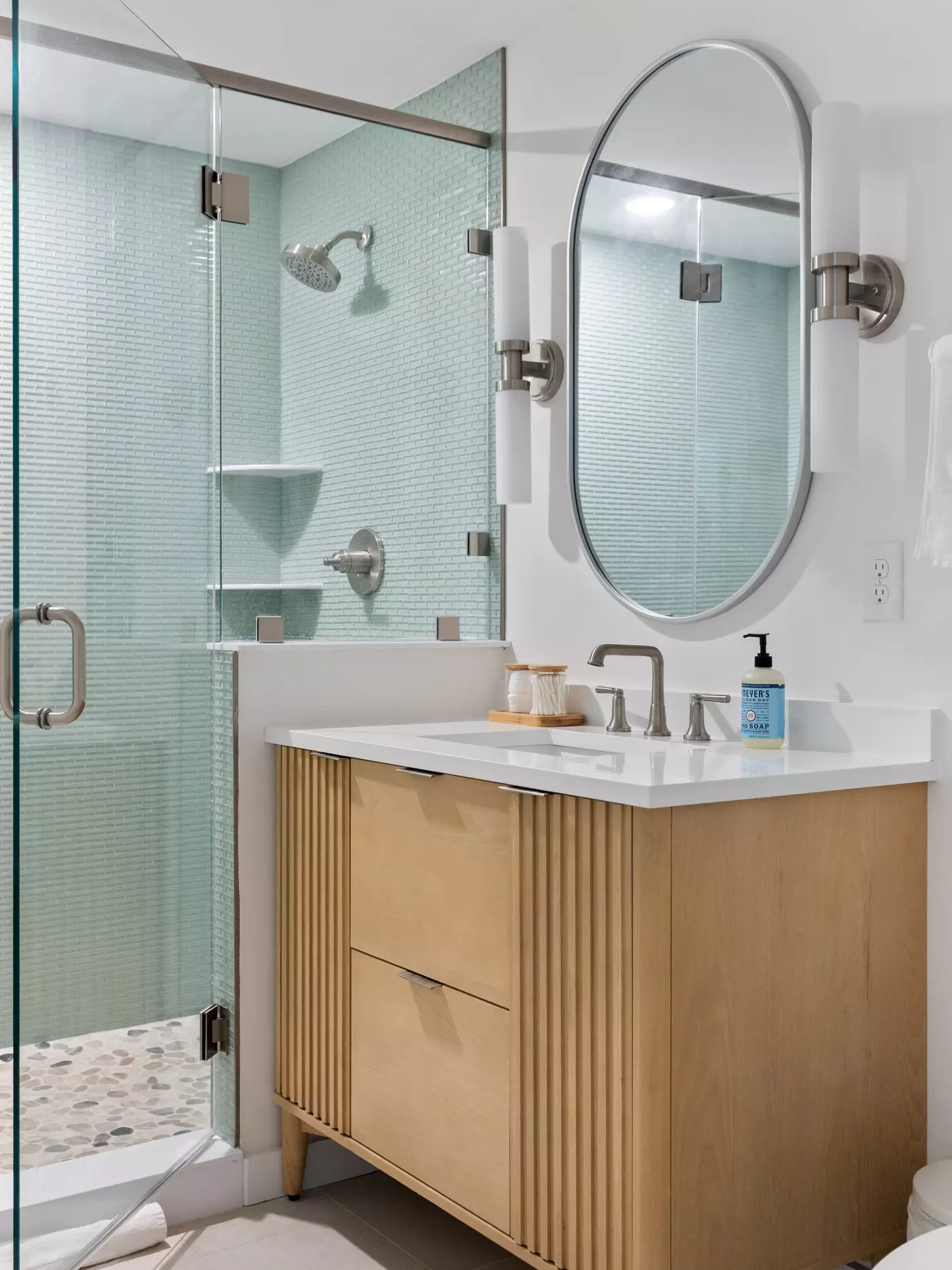 Modern bathroom with a glass shower, wooden vanity, sink, and oval mirror.