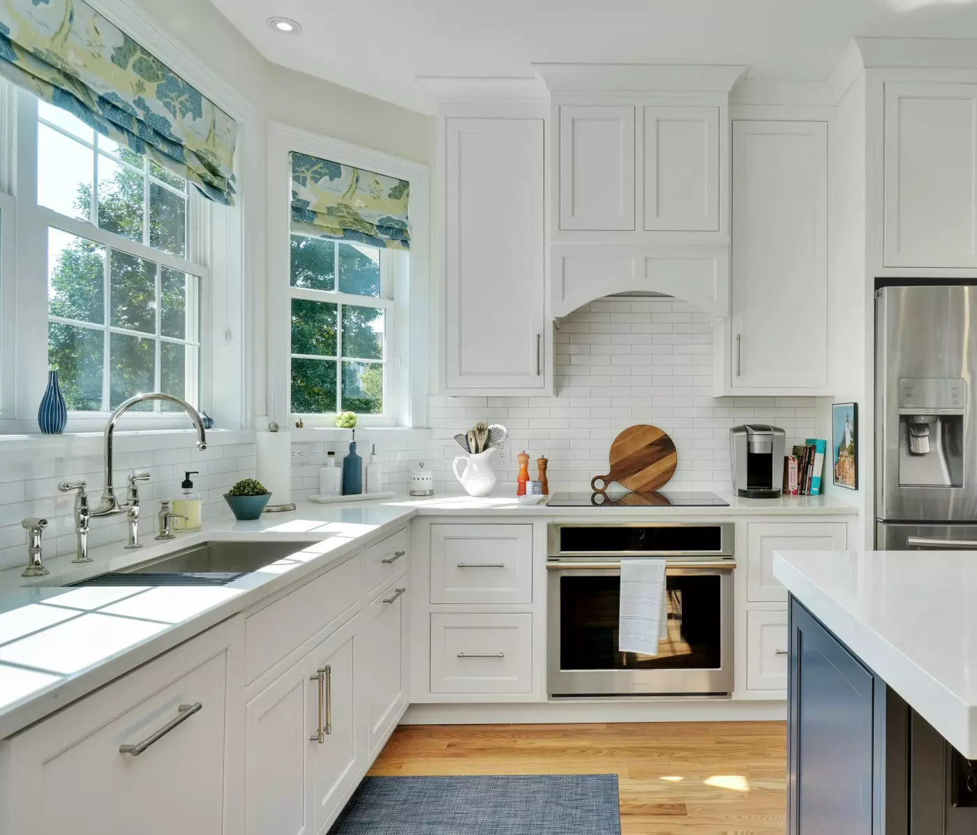 Bright white kitchen with blue island, stainless steel appliances, large windows, and floral Roman shades.
