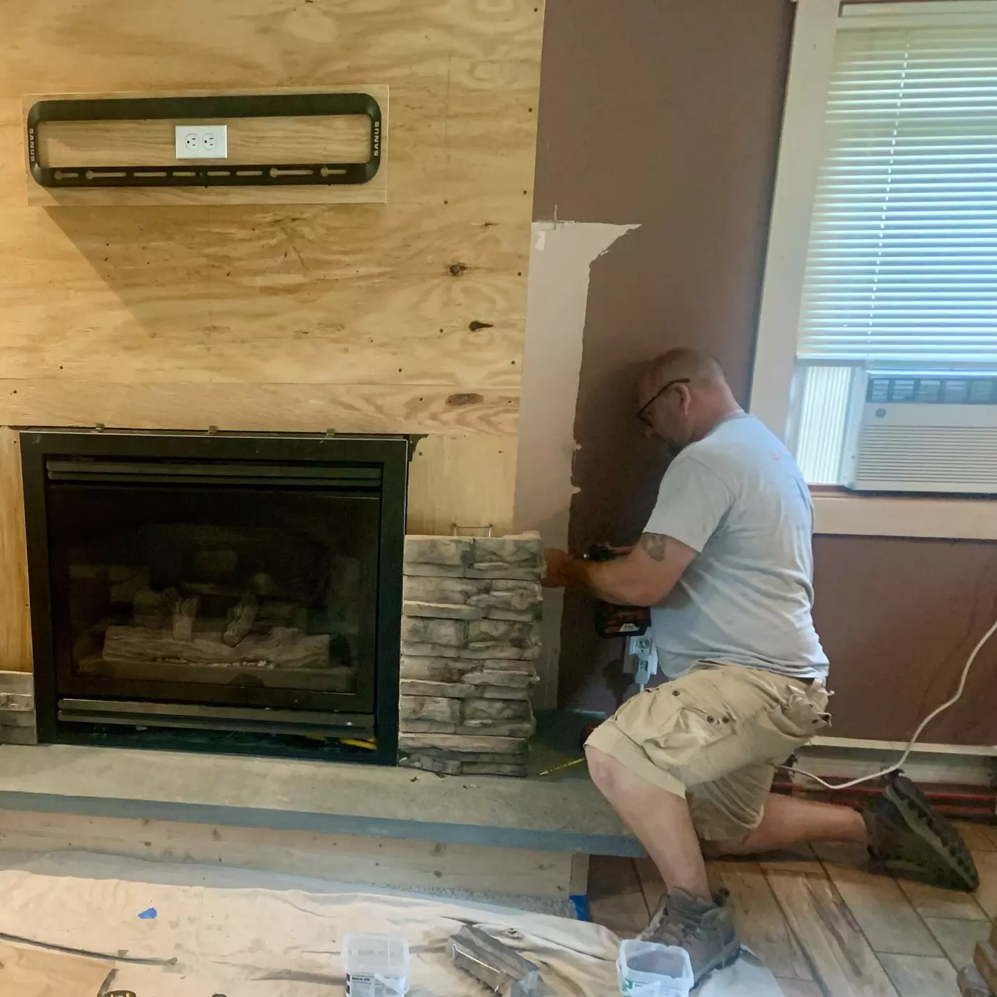 Two workers install stone around a fireplace, surrounded by construction materials.