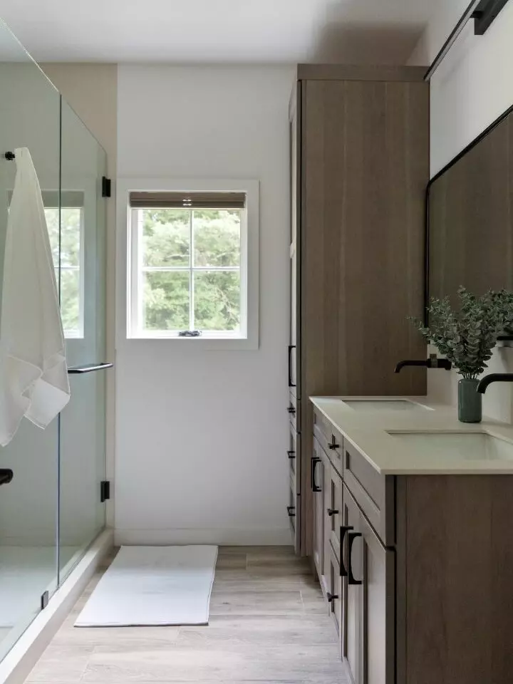 Modern bathroom with glass shower, wooden cabinets, and a window overlooking greenery.