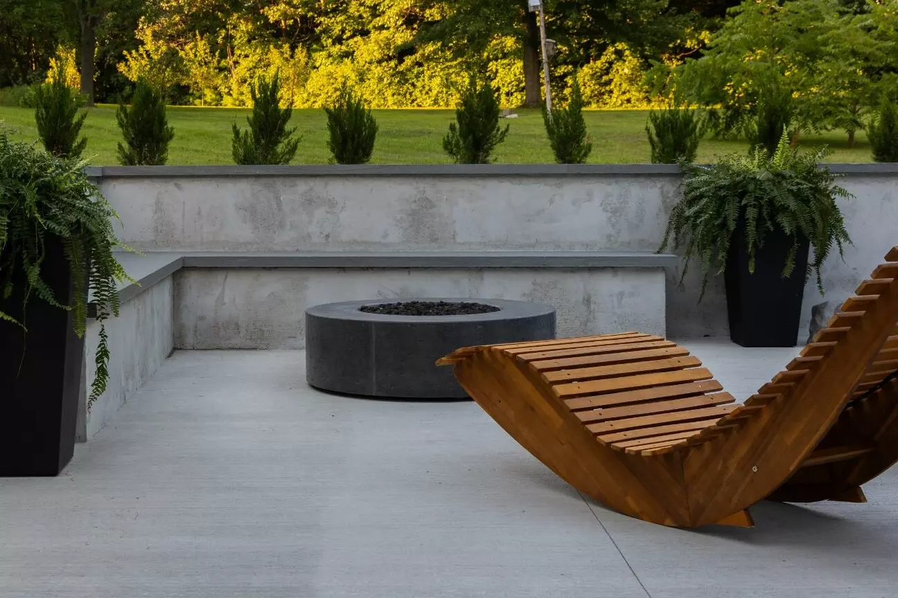Outdoor patio with wooden lounge chair, fire pit, and lush greenery.