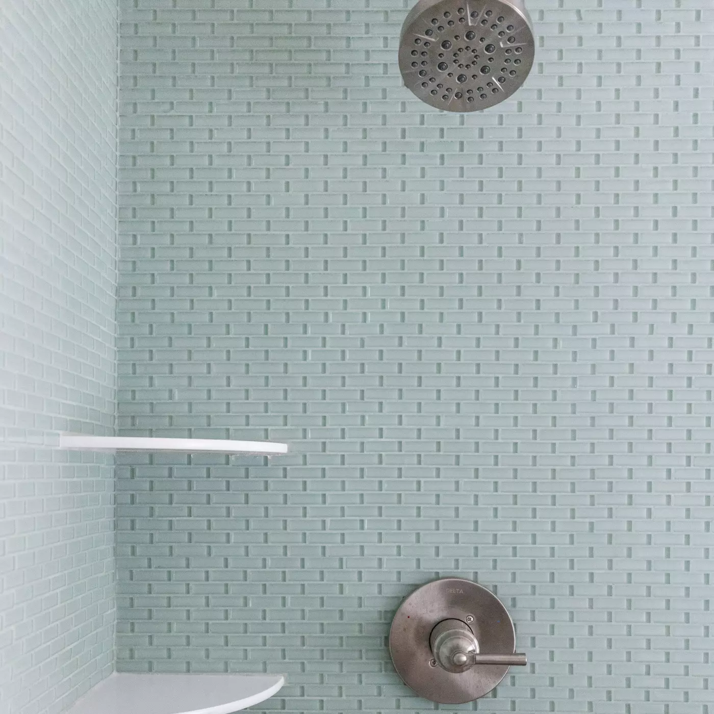 Showerhead and controls against light green textured tile with two shelves.