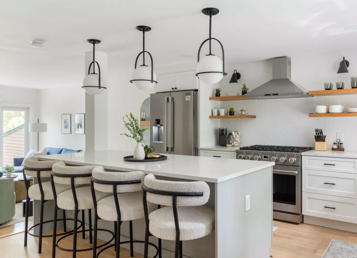 Spacious white kitchen with quartz island, black-and-white pendant lights, herringbone backsplash, and stainless steel appliances.