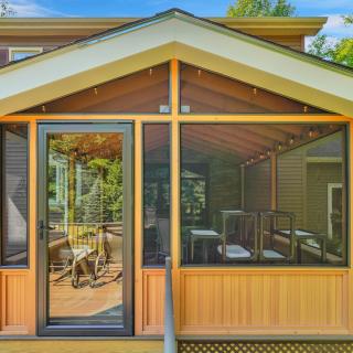 Screened porch with wooden framing and chairs, surrounded by greenery.